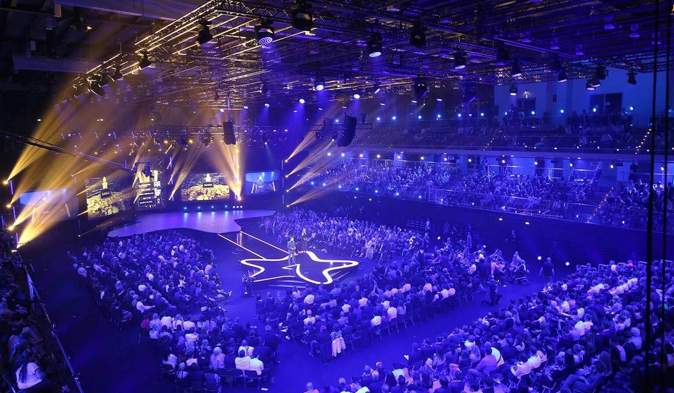Im Innenraum ist eine sternförmige Bühne aufgebaut. Die Bühne ist umgeben von Zuschauer:innen. Der Raum ist blau beleuchtet. Von der Bühne strahlen gelbe Scheinwerfer durch den Raum. 